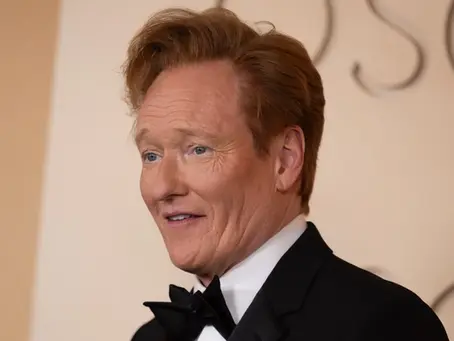 Moderator und Komiker Conan O’Brien bei einem öffentlichen Auftritt im schwarzen Smoking mit Fliege vor einer beigefarbenen Oscar-Kulisse.