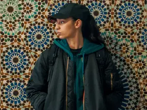 Szene aus „Die jüngste Tochter“: Fatima trägt Kappe und Jacke und steht vor einer kunstvoll gemusterten Mosaikwand, den Blick zur Seite gerichtet.