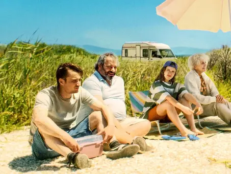 Die dysfunktionale Reisegruppe aus „Bon Voyage – Bis hierher und noch weiter“ sitzt gemeinsam an einem sonnigen Strand vor einem Wohnmobil. Unter einem Sonnenschirm verbringen Marie, Bruno, Anna und Rudy einen selten harmonischen Moment während ihres Roadtrips Richtung Schweiz.