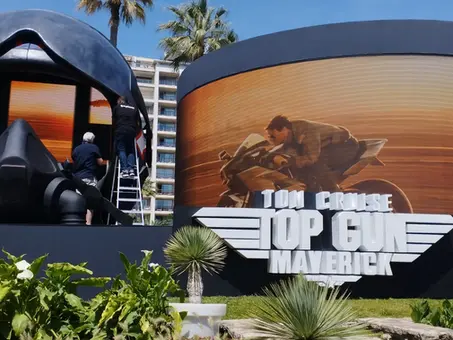 Ein großes Promo-Display für den Film „Top Gun: Maverick“ im Freien, bestehend aus einer riesigen Helm-Installation und einer gebogenen Leinwand mit einem Szenenmotiv von Tom Cruise auf einem Motorrad. Im Vordergrund stehen Pflanzen, im Hintergrund sind Palmen und Gebäude zu sehen.
