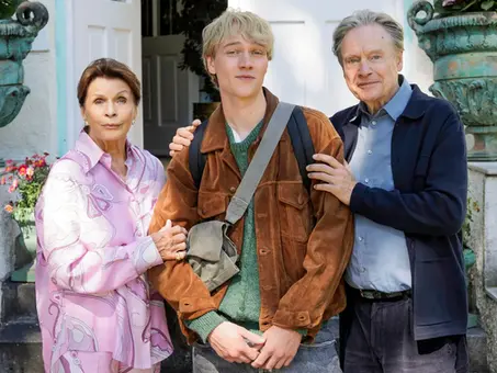 Szenenbild aus „Ach, diese Lücke, diese entsetzliche Lücke“: Senta Berger, Bruno Alexander und Michael Wittenborn stehen gemeinsam vor dem Eingang einer Villa. Berger trägt eine rosafarbene Bluse und hält den Arm von Alexander, der mit brauner Jacke und Umhängetasche in der Mitte steht. Wittenborn legt ihm von der anderen Seite die Hand auf die Schulter.