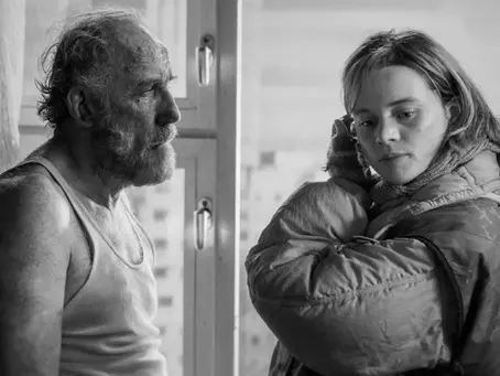 Schwarz-weiß-Aufnahme aus „Sie glauben an Engel, Herr Drowak?“: Karl Markovics als der verwahrloste Hugo Drowak steht im Unterhemd in einer kargen Wohnung und blickt zur Seite, während Luna Wedler als Lena in dicker Jacke und Schal neben ihm steht und nachdenklich aus dem Fenster schaut.