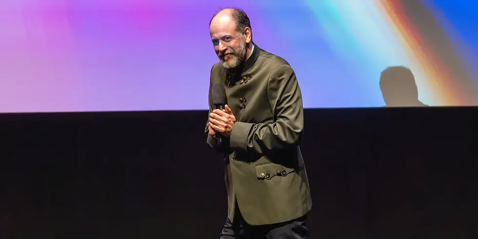 Luca Guadagnino steht auf einer Bühne vor einer großflächig farbig beleuchteten Leinwand, hält die Hände vor der Brust zusammen und blickt ins Publikum.