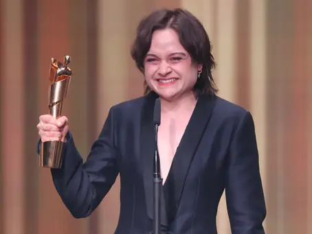 Schauspielerin Lena Urzendowsky hält beim Deutschen Filmpreis 2026 die goldene Lola-Trophäe in die Höhe und lächelt emotional auf der Bühne. Urzendowsky wurde für ihre Rolle in „In die Sonne schauen“ ausgezeichnet. Im Hintergrund sind die warm beleuchteten Vorhänge der Verleihung zu sehen.
