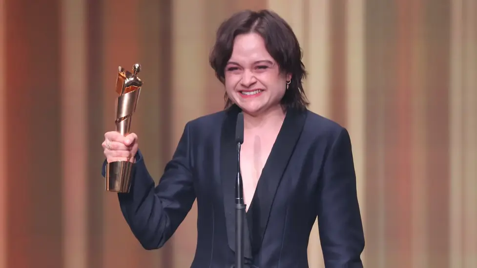 Schauspielerin Lena Urzendowsky hält beim Deutschen Filmpreis 2026 die goldene Lola-Trophäe in die Höhe und lächelt emotional auf der Bühne. Urzendowsky wurde für ihre Rolle in „In die Sonne schauen“ ausgezeichnet. Im Hintergrund sind die warm beleuchteten Vorhänge der Verleihung zu sehen.