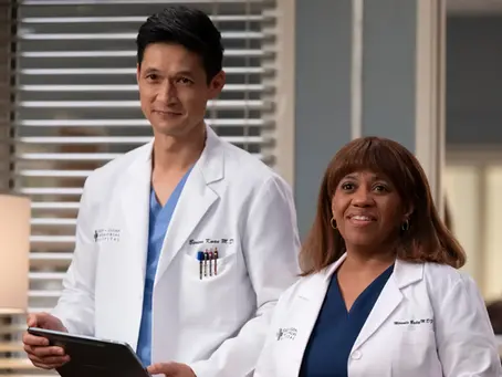 Szenenbild aus „Grey’s Anatomy“: Dr. Benson „Blue“ Kwan, gespielt von Harry Shum Jr., und Miranda Bailey, gespielt von Chandra Wilson, stehen in einem Krankenhausraum des Grey Sloan Memorial Hospital. Beide tragen weiße Arztkittel über blauer Krankenhauskleidung und blicken in die Kamera. Im Hintergrund sind Jalousien, eine Lampe sowie verschwommen weitere Personen im Krankenhaus zu sehen.