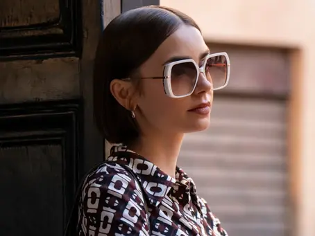 Eine junge Frau mit großen, eckigen Sonnenbrillen lehnt an einer dunklen Holztür und blickt nachdenklich zur Seite. Sie trägt ein gemustertes Oberteil, im Hintergrund ist eine unscharfe Straßenszene zu sehen. Szene aus der Netflix-Serie „Emily in Paris“.