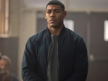 Szenenbild aus der HBO-Serie „Lanterns“: Aaron Pierre als Green-Lantern-Figur John Stewart steht in einem Innenraum mit ernster Miene vor mehreren Personen im Hintergrund und trägt eine dunkle Jacke über einem grauen Shirt.