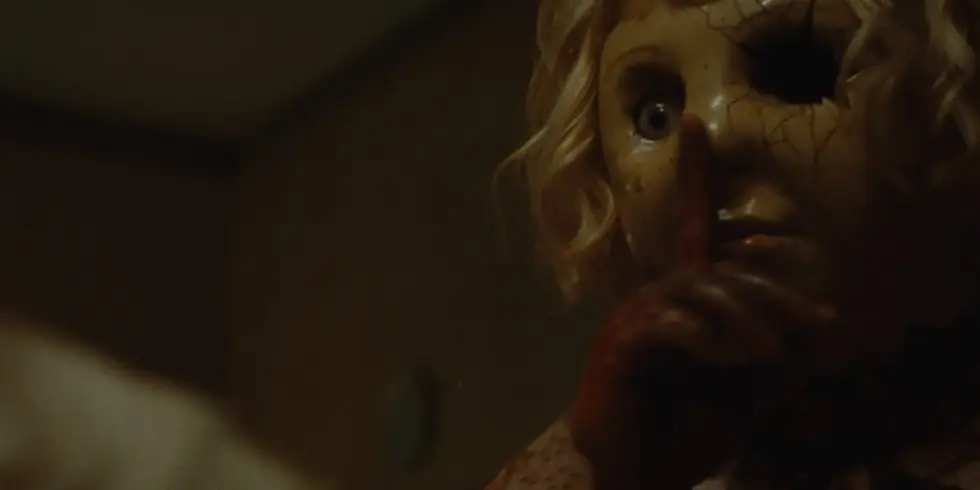 Die maskierte Killerfigur Dolly aus dem Horrorfilm „Dolly“ beobachtet ihr Opfer aus dem Schatten eines schwach beleuchteten Hauses.