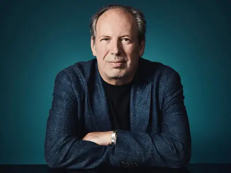 Porträt von Hans Zimmer: Der Filmkomponist sitzt vor dunklem Hintergrund, trägt ein dunkles Sakko über einem schwarzen Shirt und blickt frontal in die Kamera. Seine Arme sind locker vor dem Körper verschränkt.
