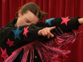 „Sunny Dancer“: Eine Jugendliche tanzt vor einem roten Vorhang in einer schwarzen Jacke mit bunten Sternen.
