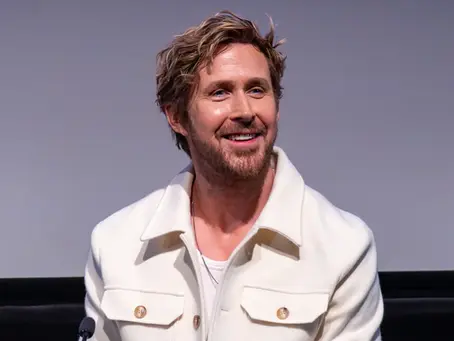Ryan Gosling sitzt lächelnd in einer weißen Jacke bei einer Veranstaltung vor einer grauen Leinwand.