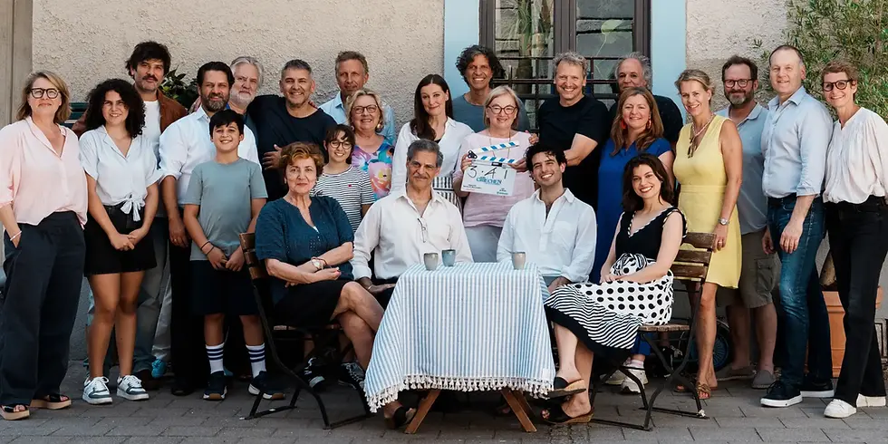 Gruppenfoto von Cast und Team des Films „Beim Griechen“. Vorn sitzen Despina Pajanou, Rick Kavanian, Nicolas Sidiropulos und Violeta Lupis an einem Tisch, dahinter steht das weitere Ensemble und Filmteam. In der Mitte wird eine Filmklappe in die Kamera gehalten.