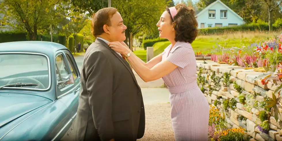Szenenbild aus „Die Progressiven Nostalgiker“: Hélène und Michel stehen im Freien neben einem Auto der 1950er-Jahre, sie richtet ihm den Kragen – Moment ehelicher Nähe vor der Zeitreise.