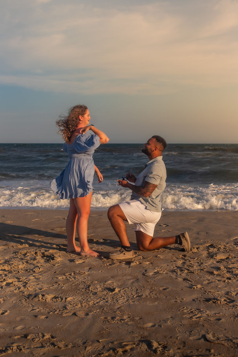 Man kneels on a sandy beach to propose to a smiling woman in a blue dress, with waves and a sunset sky behind them.