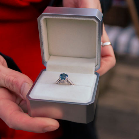 Hands holding an open ring box with a blue gemstone ring, blurred background, suggesting a proposal or gift moment.