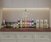 A modern home café setup displayed on a dark wooden countertop against a cream marble backsplash with warm under-cabinet lighting. A glowing white script sign reading "At Home Café" sits beneath a green Sims plumbob. The counter is neatly lined with coffee and matcha products, cocoa powder, fruit refreshers, flavoured syrup bottles (including chocolate, vanilla, caramel, mango, strawberry, lavender, and blueberry), plant-based milk cartons (almond, oat, and coconut), canned matcha lattes, and a pink handheld milk frother. White shaker-style cabinets frame the scene, creating a clean, cosy café aesthetic.