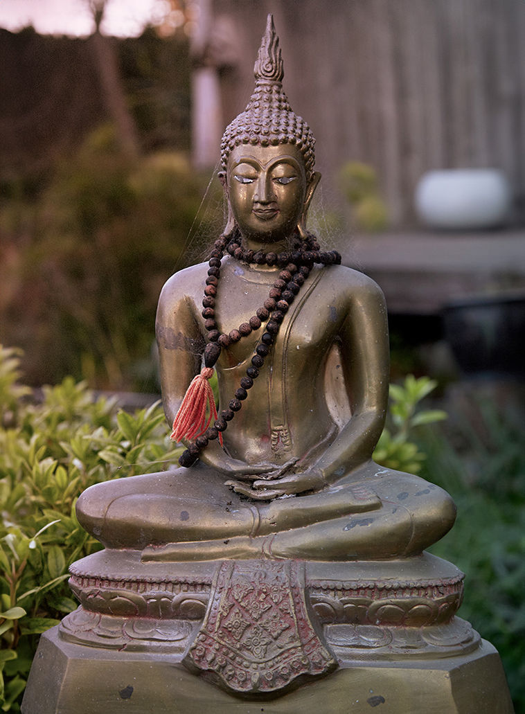 Bronze Buddha statue with mala beads and a red tassel, seated in meditation in an outdoor garden at East West’s Oakland location, symbolising mindfulness and healing presence.
