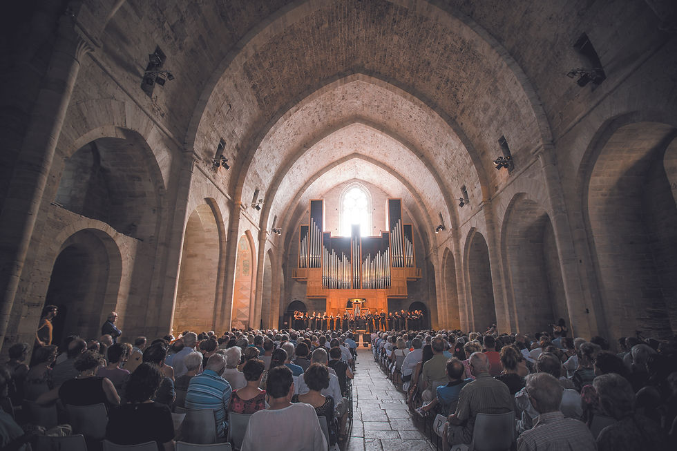 Concert choral dans une église voûtée en pierre de l'abbaye de Sylvanès public assis face à un grand orgue éclairé par une fenêtre centrale.