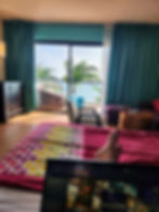 Person relaxing in a colorful hotel room with ocean view; polka dot bedspread, laptop open, palm trees outside.