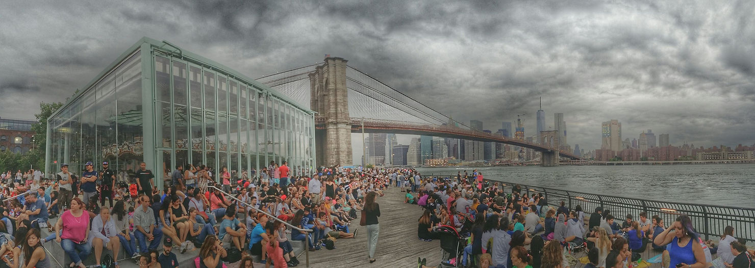 Crowd gathers by a carousel near Brooklyn Bridge under cloudy skies. People sit on steps and boardwalk, with New York City skyline in the background.