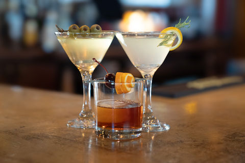Three cocktails with olives, lemon, and orange garnish on a bar