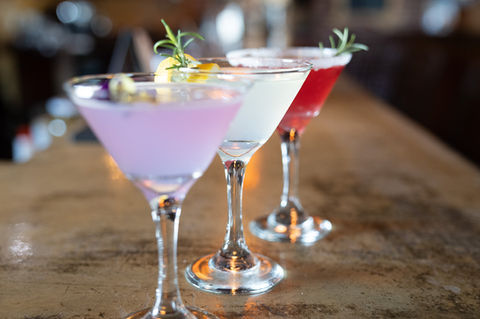 Three colorful cocktails garnished with herbs sit on a wooden bar surface.