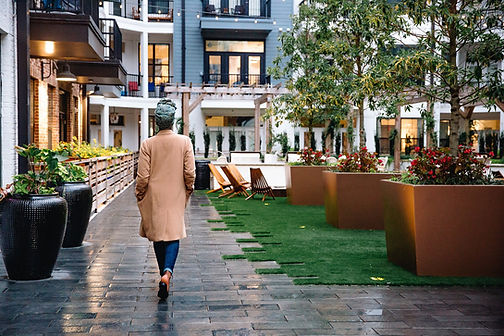 Diana is walking outside, away from the camera. She is wearing a brown wool jacket and blue jeans. Trees, plants, and a building are in the frame.