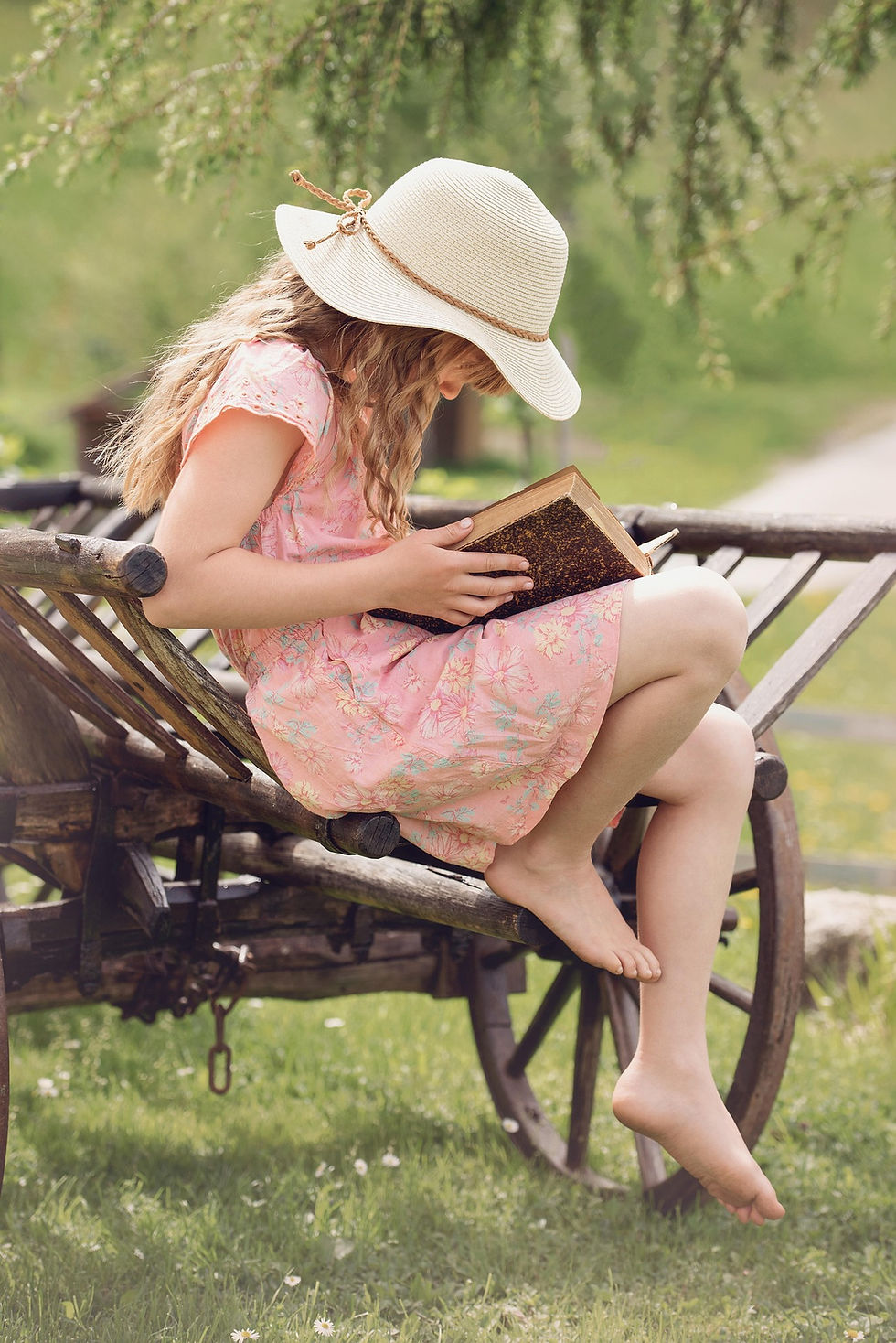 A girl with a hat on sitting in a summer garden reading a book