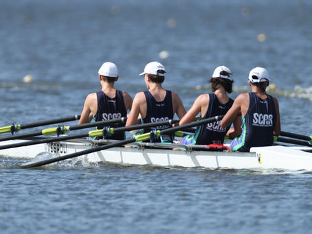 A SCAR boys quad rows together during a regatta, wearing matching navy racing uniforms and white hats. The crew powers across the water in sync with their oars moving evenly through the course.