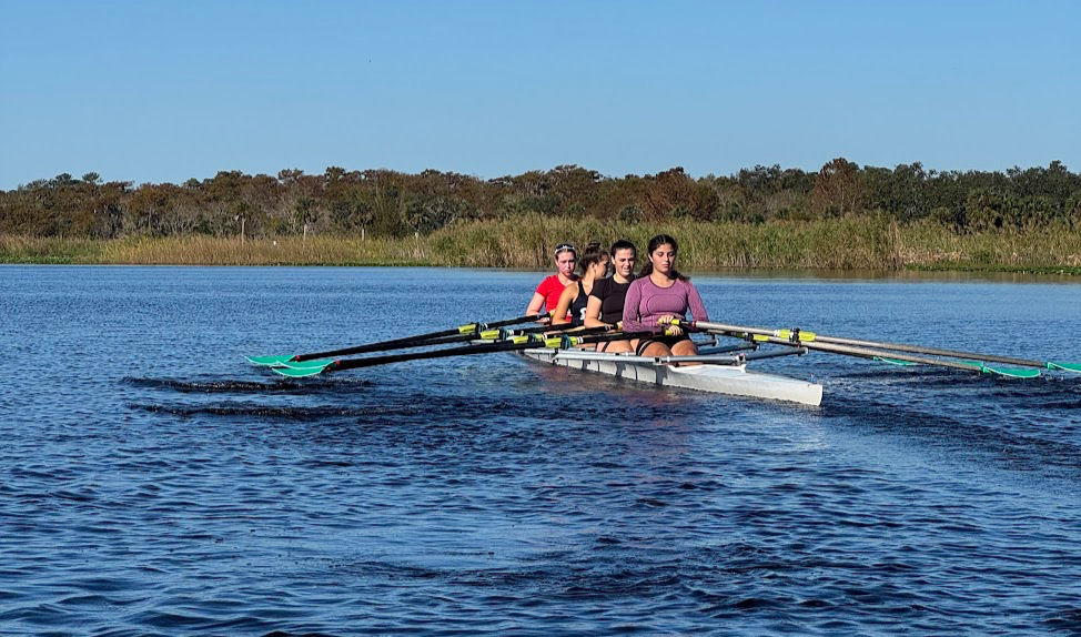 A SCAR women’s rowing quad moves across Lake Jesup during practice on a clear afternoon. The four athletes row in sync in a sweep boat with green-bladed oars, surrounded by calm blue water and marshland in the background.