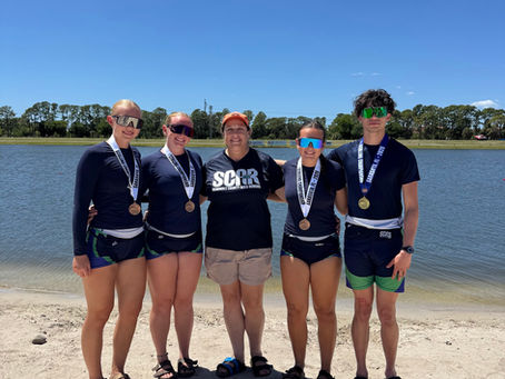 SCAR rowers and Head Coach Michelle Hill pose with medals on the shoreline after the FSRA Sculling State Championships. Athletes from Seminole, Lyman, and Lake Mary High Schools stand together in racing uniforms under a bright blue sky.