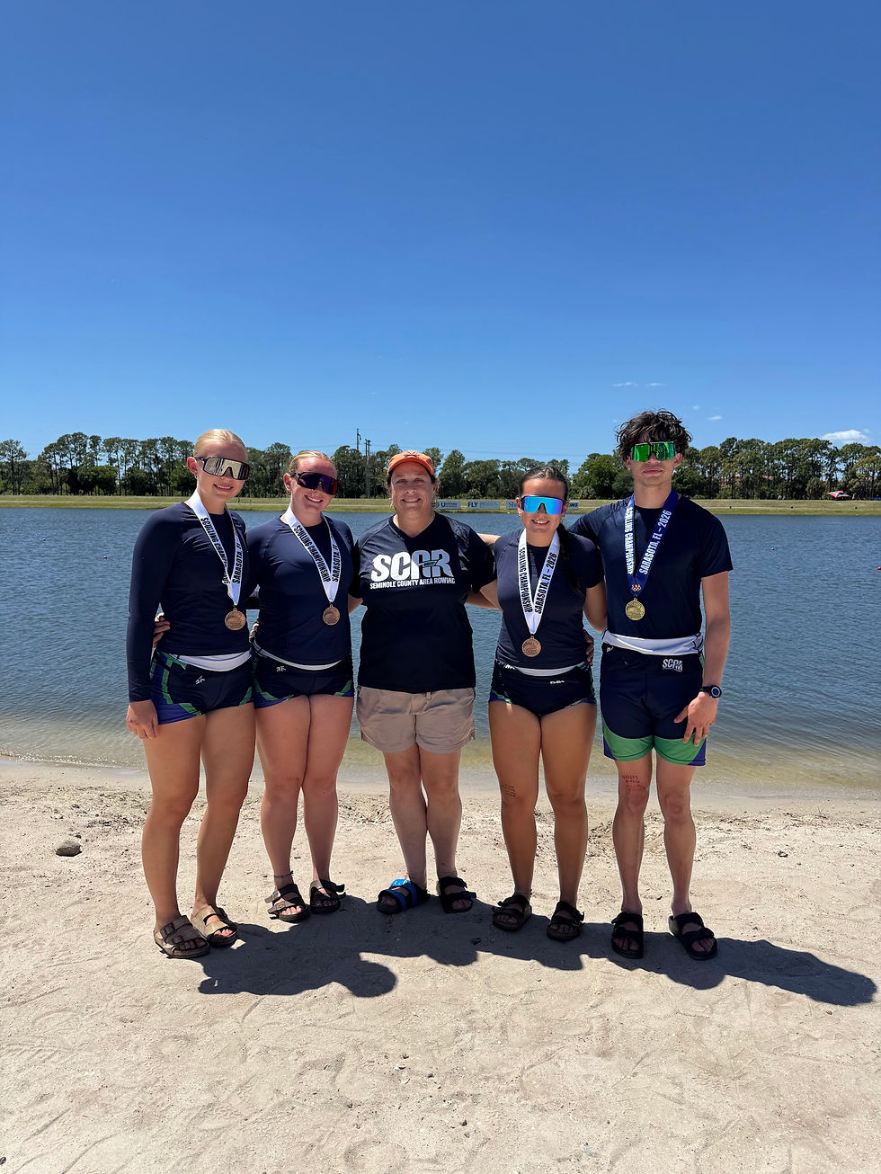 SCAR rowers and Head Coach Michelle Hill pose with medals on the shoreline after the FSRA Sculling State Championships. Athletes from Seminole, Lyman, and Lake Mary High Schools stand together in racing uniforms under a bright blue sky.