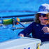 SCAR coxswain wearing a navy team shirt, headset, sunglasses, and SCAR hat while steering a rowing shell in Central Florida during a race. The coxswain focuses on guiding the crew as rowers and oars move through calm blue water in the background.
