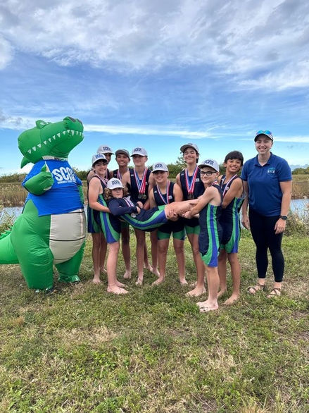 Development rowers gather with SCAR’s inflatable alligator mascot and Coach Alex after a regatta, smiling and celebrating their accomplishments.