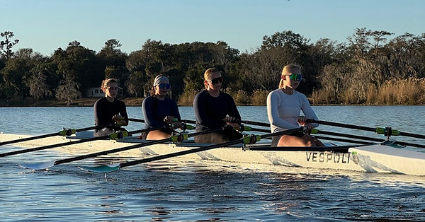 Four rowers paddle together in a Vespoli shell during practice on calm water.