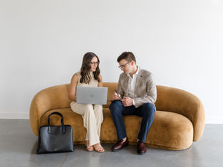A professional man and woman sitting on a sofa with a computer having a discussion.
