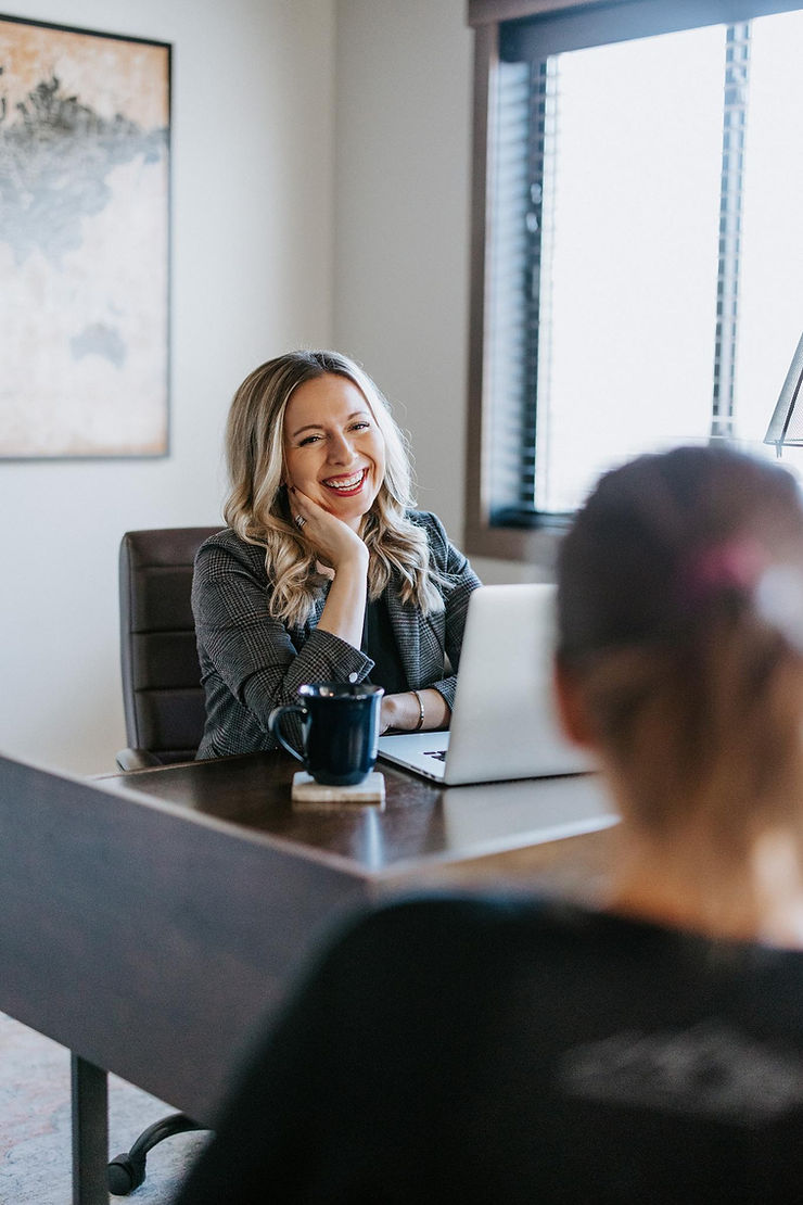 Smiling woman in an office, seated at a desk with a laptop and mug. Another person is partially visible. Bright and professional mood.