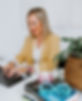Close-up of a desk with plants, a pile of books topped with turquoise-framed glasses and a mini disco ball candle, and Liz Carlson’s hands—wearing bracelets and rings—typing on a notebook blurred in the background.