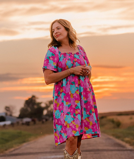 Emily standing in pink and blue floral dress in front of a sunset orange sky
