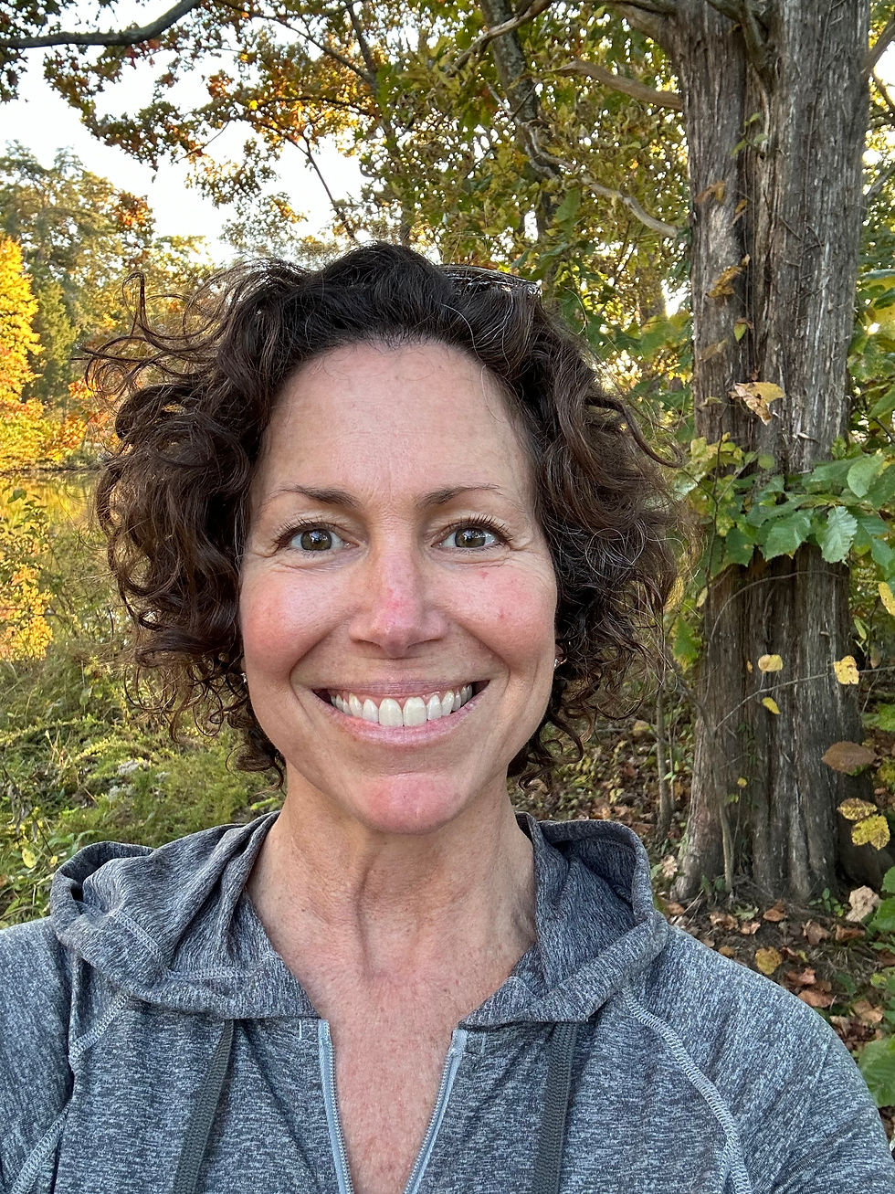 A smiling person in a gray hoodie stands outdoors. Behind them are trees with green and yellow leaves, suggesting a fall setting.