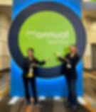 Two women smile and gesture towards a large sign reading "AAD Annual Meeting" on a green and blue backdrop. Indoor setting with colorful carpet.