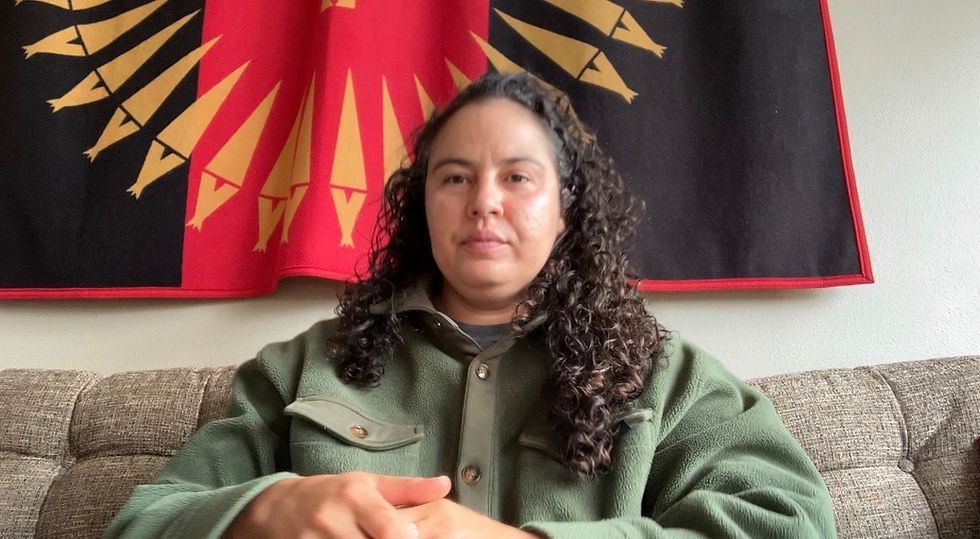 Person with curly hair sits on a couch in a green jacket, in front of a red-and-black flag with yellow arrow designs.