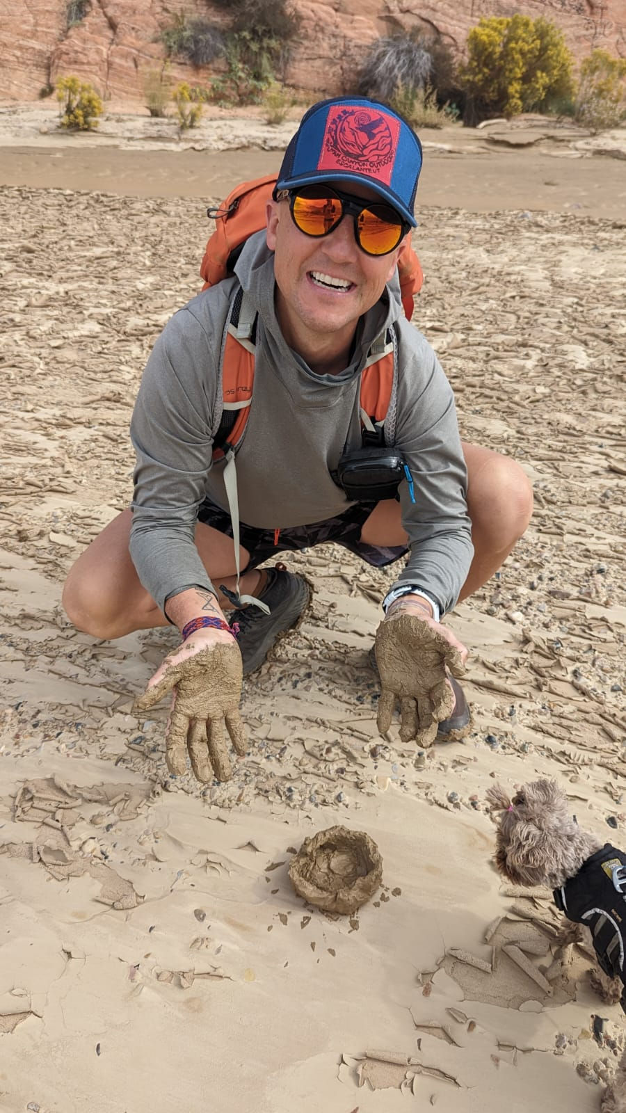 Man in hat and sunglasses kneeling on sandy ground with muddy gloves, next to a dog. Rocky background, cheerful expression.