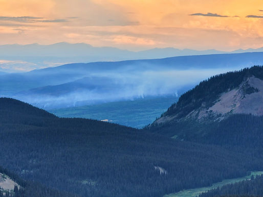 Mountainous landscape with dark green forests, layers of hills, and a pastel orange sunset sky. Smoke rises from the valleys, creating an ominous mood.