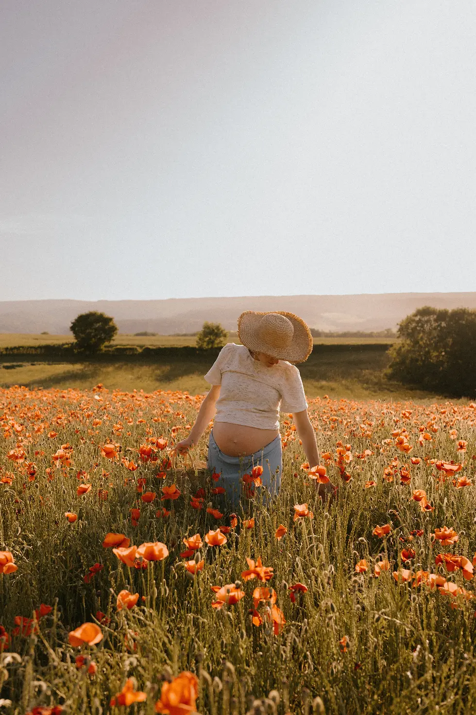 Femme enceinte au chapeau de paille dans un champ de coquelicots orange, sous un ciel clair, ambiance paisible.
