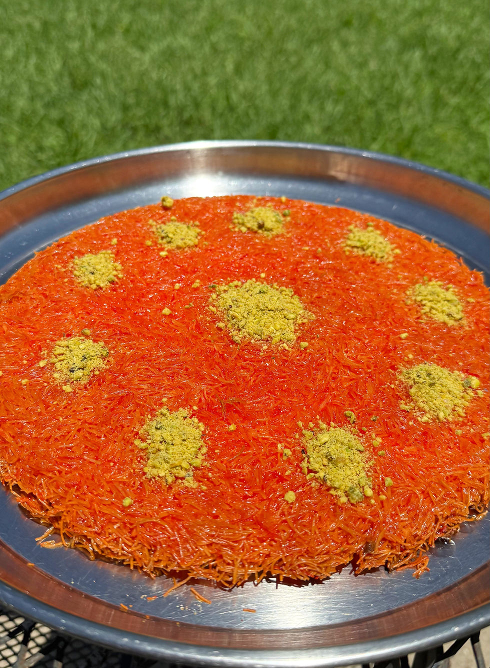 Round orange dessert topped with green pistachio bits on a metal tray, set outdoors on grass. Bright and inviting scene.