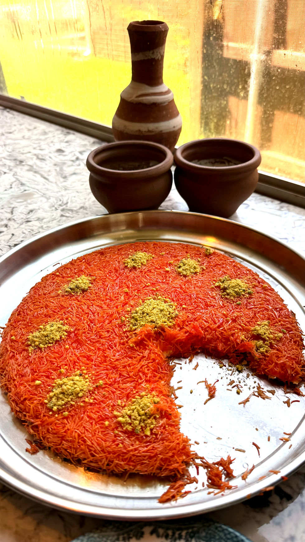 Orange dessert with green toppings on a metal tray, a slice missing. Clay pots and a jug on a windowsill background, soft lighting.