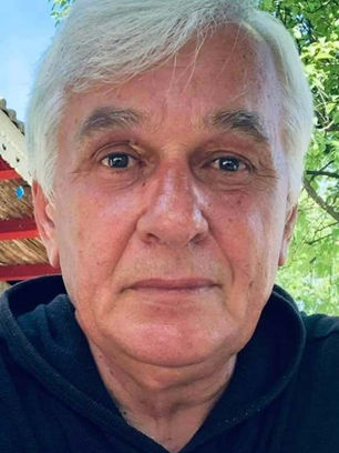Artist Marian Năstase, purtând un hanorac negru, zâmbind ușor. Fundal cu verdeață și cer senin, exprimând o stare de calm.