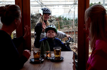 Twee kinderen zwaaien naar hun moeders met fietshelmen achter glas. Binnen staan glazen op tafel, omgeving van een serre.